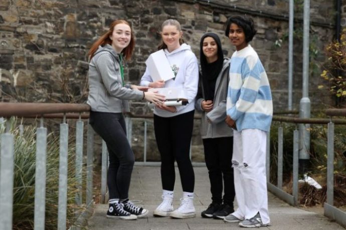 Four young people smiling at the camera standing on a bridge. They are dressed in casual clothes and one of them is holding a box.