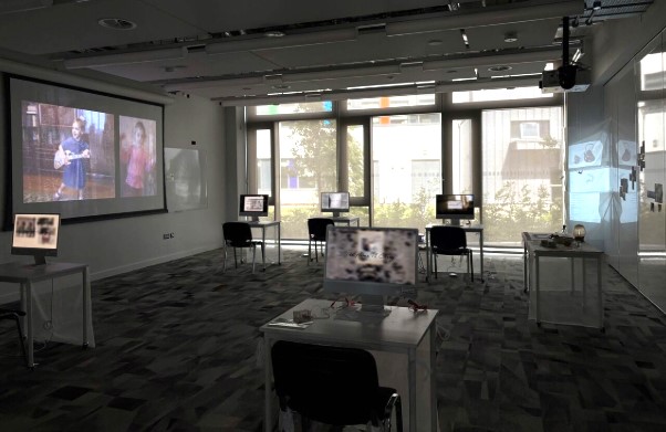 A contemporary exhibition room with a projection screen showing a video, several transparent tables with computer monitors and natural light filtering through large windows.