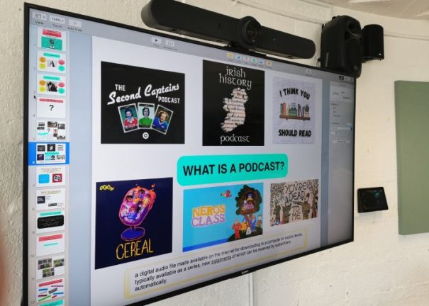 A large wall mounted screen is displaying a colorful PowerPoint presentation to help children learn about podcasting. The words 'What is a Podcast' is displayed in large black text against a bright green background.